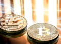 Two physical coins representing Litecoin and Bitcoin rest on a reflective surface, with abstract light patterns in the background. The Litecoin coin features the "L" logo, whilst the Bitcoin coin displays the "B" symbol. | FintechZoom