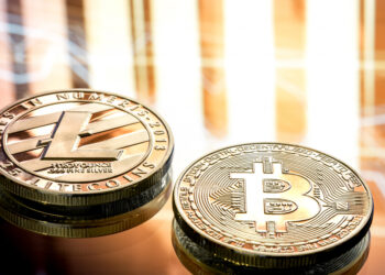 Two physical coins representing Litecoin and Bitcoin rest on a reflective surface, with abstract light patterns in the background. The Litecoin coin features the "L" logo, whilst the Bitcoin coin displays the "B" symbol. | FintechZoom