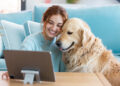 A smiling woman sits on a sofa next to a golden retriever, both looking at a tablet on a table in front of them. The cosy living room features light blue cushions in the background. | FintechZoom