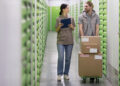 Two people walk through a storage facility corridor with green doors. One holds a clipboard, and the other pushes a trolley stacked with cardboard boxes labelled "Express." They appear to be talking and working together. | FintechZoom