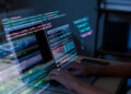 A person is typing on a laptop displaying code on multiple screens, with colourful lines of programming text visible, suggesting software development or coding work in progress. | FintechZoom