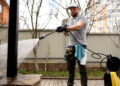 A man wearing a cap and gloves uses a pressure washer to clean an outdoor surface in a garden, with trees, a fence, and blocks of flats in the background. | FintechZoom