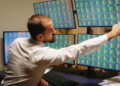 A man in a white shirt sits at a desk with multiple monitors displaying financial data and share charts, reaching to adjust one of the screens. Papers with charts rest on the desk nearby. | FintechZoom
