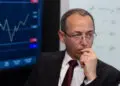 A man in a suit and red tie sits thoughtfully with his hand on his chin, in front of a screen showing a rising financial graph and a whiteboard with handwritten notes. | FintechZoom