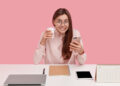A young woman with glasses, smiling and holding a coffee cup in one hand and a mobile phone in the other, sits at a white desk with a laptop, notebook, pen, tablet, and diary against a pink background. | FintechZoom
