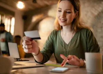 A woman sits at a desk, smiling whilst holding a credit card in one hand and using a mobile phone with the other. A laptop, cup, and notepad are on the desk in a softly lit, cosy room. | FintechZoom