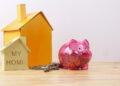A pink piggy bank filled with coins, a set of house keys, and two model houses—one labelled "MY HOME"—are arranged on a wooden surface against a plain background. | FintechZoom