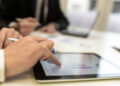 Close-up of hands holding pens and analysing a bar graph on a tablet screen, with blurred figures and a laptop in the background, suggesting a business meeting or discussion. | FintechZoom