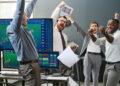 Four men in business attire celebrate excitedly in an office with computer screens displaying share charts, one man holding a headset and tablet, others raising arms, indicating a successful financial achievement. | FintechZoom