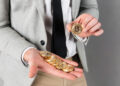 A person in business attire holds several gold-coloured Bitcoin tokens in one hand and displays one Bitcoin token in the other hand. | FintechZoom