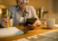A woman sitting at a desk smiles whilst looking at her mobile. She holds a yellow pencil over a notepad, with a coffee mug and computer keyboard nearby. The setting appears warm and cosy. | FintechZoom