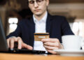 A person in a suit sitting at a table uses a laptop whilst holding a bank card. A white coffee cup is on the table in the foreground. The scene suggests online shopping or digital banking. | FintechZoom
