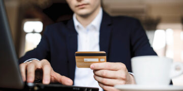 A person in a suit sitting at a table uses a laptop whilst holding a bank card. A white coffee cup is on the table in the foreground. The scene suggests online shopping or digital banking. | FintechZoom