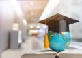 A small globe wearing a graduation cap sits on an open book, with a blurred indoor background, symbolising global education and learning. | FintechZoom