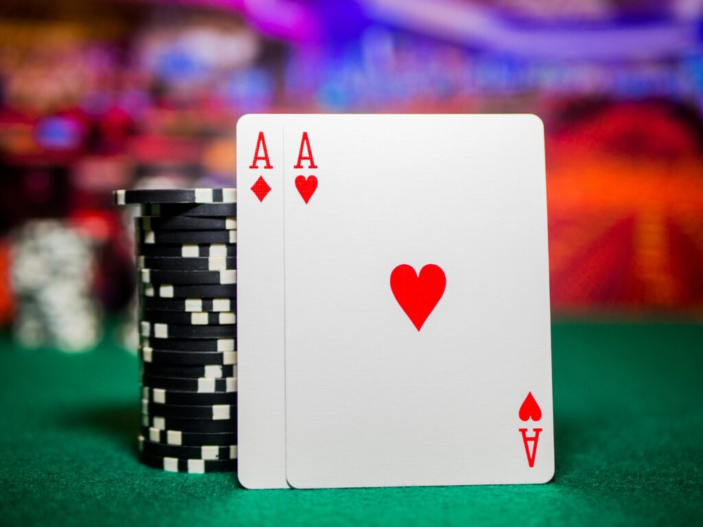 Two aces of hearts playing cards are shown leaning against a stack of black and white poker chips on a green baize table, with a colourful blurred background. | FintechZoom