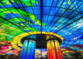 A vibrant stained glass ceiling with bold blue, green, yellow, and red colours forms a dome above two illuminated cylindrical pillars, creating a colourful and artistic indoor space. | FintechZoom