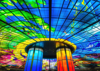 A vibrant stained glass ceiling with bold blue, green, yellow, and red colours forms a dome above two illuminated cylindrical pillars, creating a colourful and artistic indoor space. | FintechZoom
