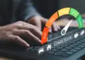 A person types on a laptop keyboard, with an illustrated credit score gauge overlay showing levels from "Poor" to "Excellent," and the indicator points to "Good." Icons of a car, house, and pound sign are also visible. | FintechZoom