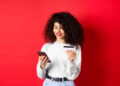 A woman with curly hair smiles whilst looking at her mobile and holding a credit card, standing against a bright red background. She is wearing a light grey jumper and jeans. | FintechZoom