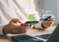 A person holding a mobile phone and a credit card, making an online payment at a desk with a laptop and calculator. A digital "Payment" graphic with a security tick is overlaid on the image. | FintechZoom