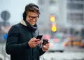 A young man wearing glasses and headphones smiles while looking at his mobile. He is holding a coffee cup and standing outside on a city street with blurred traffic lights and cars in the background. | FintechZoom