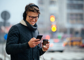 A young man wearing glasses and headphones smiles while looking at his mobile. He is holding a coffee cup and standing outside on a city street with blurred traffic lights and cars in the background. | FintechZoom