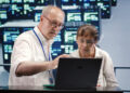 Two older adults, a man and a woman, both wearing glasses and white shirts, look intently at a laptop screen. The man is pointing at the screen, and a wall of computer monitors is visible in the background. | FintechZoom