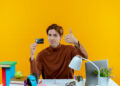 A young person sitting at a desk holds a credit card in one hand and gives a thumbs-up with the other. The desk has books, a lamp, and stationery, and the background is bright yellow. | FintechZoom