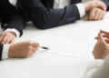 Three people in business attire sit at a table; one points at a document with a pen whilst discussing its contents with the others. | FintechZoom
