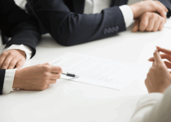 Three people in business attire sit at a table; one points at a document with a pen whilst discussing its contents with the others. | FintechZoom