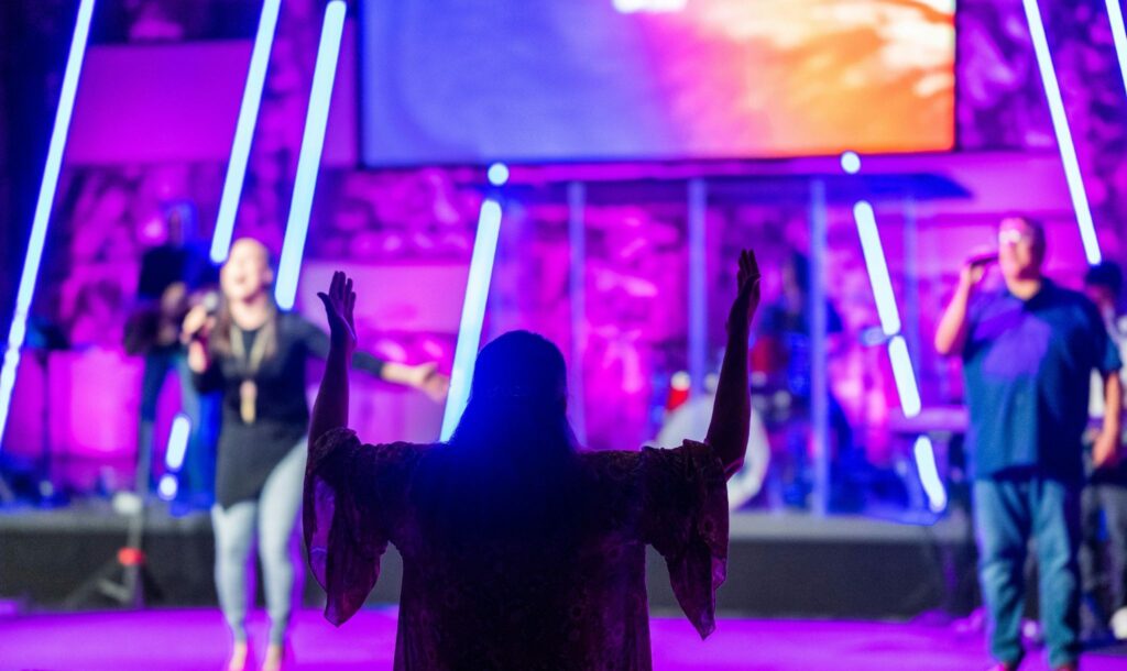 A person with raised arms stands in a brightly lit room with purple and blue lighting, facing a stage where musicians and singers perform in front of a screen. | FintechZoom