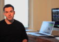 Tim Sykes sits at a desk with a laptop and a large monitor displaying share charts and graphs. A window is in the background, letting in natural light. The person appears to be working or explaining something. | FintechZoom