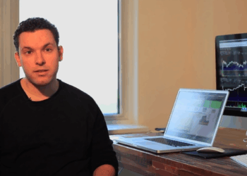 Tim Sykes sits at a desk with a laptop and a large monitor displaying share charts and graphs. A window is in the background, letting in natural light. The person appears to be working or explaining something. | FintechZoom