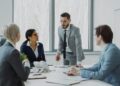 Four business professionals in suits have a serious discussion around a conference table in a modern office. One man is standing and speaking, while the others sit and listen attentively. Large windows provide natural light. | FintechZoom