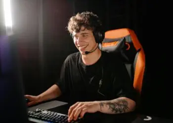 A young man with a headset smiles whilst sitting in an orange gaming chair, using a computer keyboard and mouse in a dark room. | FintechZoom