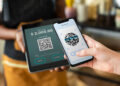 A person holds a mobile scanning a QR code on a tablet screen displaying a £2,000 payment. The background is blurred, showing an indoor setting. | FintechZoom