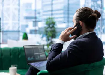 A man in a suit sits on a green settee, holding a mobile to his ear and looking at a laptop displaying graphs, in a modern office with large windows and city views. | FintechZoom