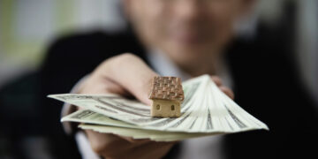 A person holds out a small model house placed on top of a fan of hundred-dollar notes, with the background blurred and the face out of focus. | FintechZoom