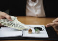 A person in a suit holds a fan of U.S. dollar notes over an open notebook with two small house models and a set of keys on top, suggesting a property transaction or investment. | FintechZoom