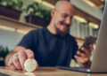 A man holding a physical Bitcoin coin at a desk, smiling whilst looking at his mobile, with a laptop nearby in a modern indoor setting. | FintechZoom