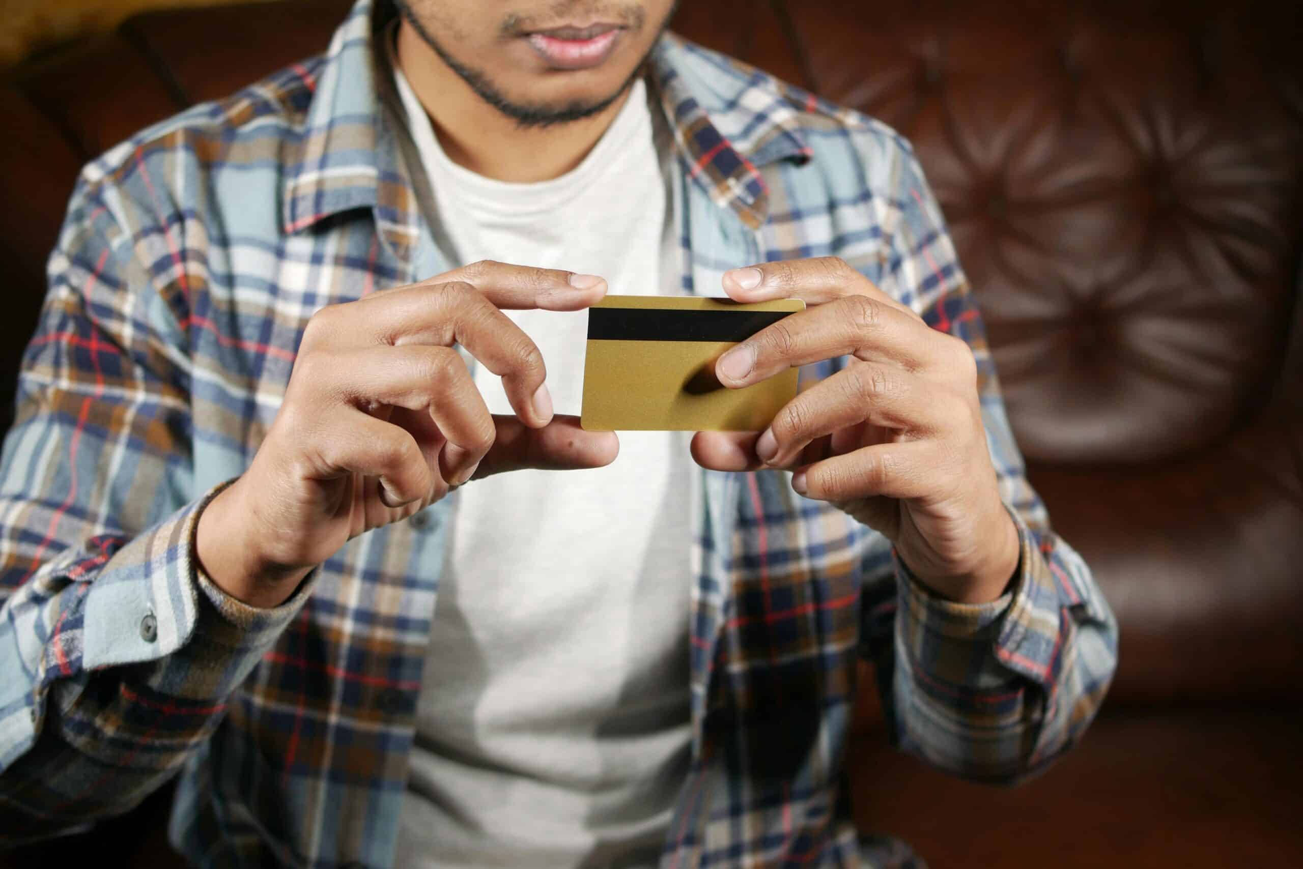 A person wearing a checked shirt and white t-shirt sits on a brown leather sofa, holding a gold credit card with both hands, focusing on the card. | FintechZoom