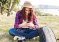 A young woman wearing a hat and checked shirt sits on the grass, smiling at her mobile. She is sitting cross-legged beside a rucksack and a camera, with trees and a river in the background. | FintechZoom