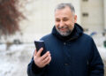 A smiling man with grey hair and a beard, wearing a dark winter coat, stands outdoors in a snowy setting while looking at his mobile phone. | FintechZoom