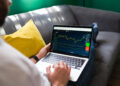 A person sitting on a sofa with a yellow cushion, using a laptop displaying a colourful stock market or trading chart with various graphs and indicators. | FintechZoom