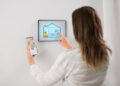 A woman controls a smart home system using a wall-mounted touchscreen panel and a mobile phone, adjusting settings and icons in a modern, bright room. | FintechZoom