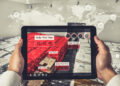 A person holds a tablet displaying augmented reality data on order pick times over stacks of packages in a warehouse, with a world map and digital icons overlaid in the background. | FintechZoom