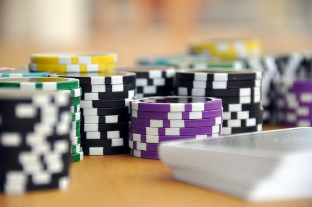Stacks of colourful poker chips and a pack of playing cards are arranged on a wooden surface, suggesting a scene from a poker game or casino setting. | FintechZoom