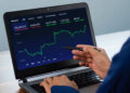 A person holding a pen points at a laptop screen displaying a financial chart with stock market data and indices in various categories such as US, Futures, and Global. | FintechZoom
