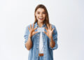 A smiling woman in a denim shirt stands against a plain background, holding a blank white card with both hands and looking excitedly at the camera. | FintechZoom