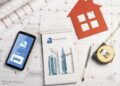 A smartphone with a property app, a notepad with building sketches, a pen, a tape measure, and a red house-shaped cut-out sit on architectural plans. | FintechZoom